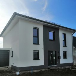 TomaszMajewski - Nowoczesny dom dwukondygnacyjny z elewacją w kolorze białym i szarym, czarnymi oknami i drzwiami, widok z zewnątrz w słoneczny dzień.