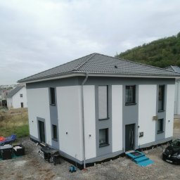 TomaszMajewski - Nowo wybudowany dom jednorodzinny z elewacją w odcieniach bieli i szarości, z widocznym fragmentem dachu pokrytego szarą dachówką, na posesji z gruzem i materiałami budowlanymi, z samochodem...