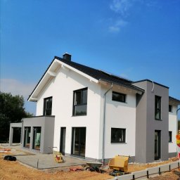 TomaszMajewski - Nowoczesny dom jednorodzinny z biało-szarą elewacją w trakcie wykończenia terenu, widoczne narzędzia budowlane i ubijarka wibracyjna.