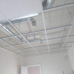 TomaszMajewski - Montaż sufitu podwieszanego kasetonowego z widocznymi instalacjami wentylacyjnymi i kablami w jasnym pomieszczeniu.