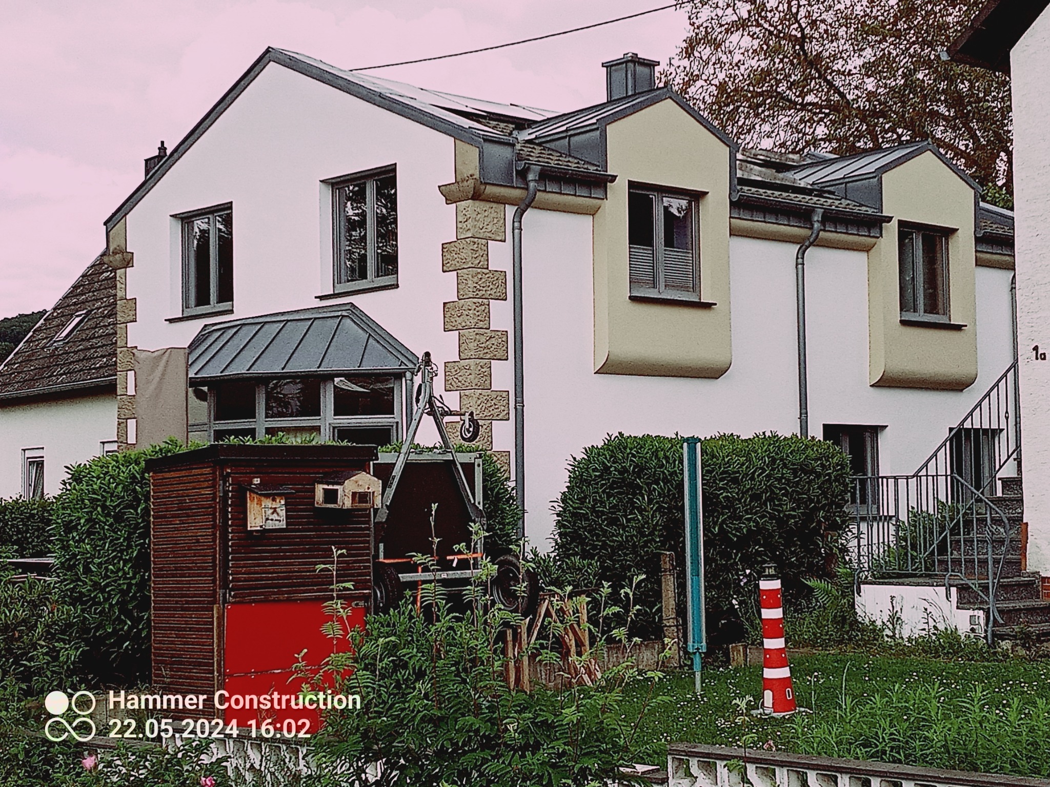 Dwupiętrowy dom z białą elewacją, elementami wykończeniowymi w odcieniach beżu i szarości, widoczne panele słoneczne na dachu, otoczony zielenią, z małą czerwoną budką i ozdobną latarnią morską...