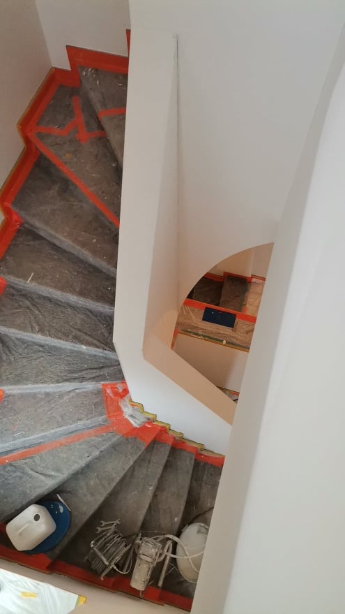 Klatka schodowa zabezpieczona folią i taśmą malarską przed rozpoczęciem prac wykończeniowych, widoczne narzędzia malarskie i drabina na schodach.