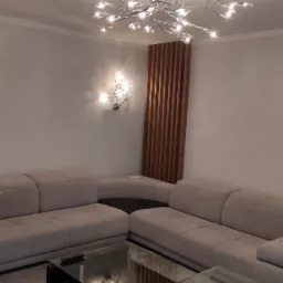 Jasny salon z narożną sofą, szklanym stolikiem kawowym i designerską lampą sufitową w kształcie gałęzi z kryształkami, oświetlona drewniana ścianka dekoracyjna w tle.