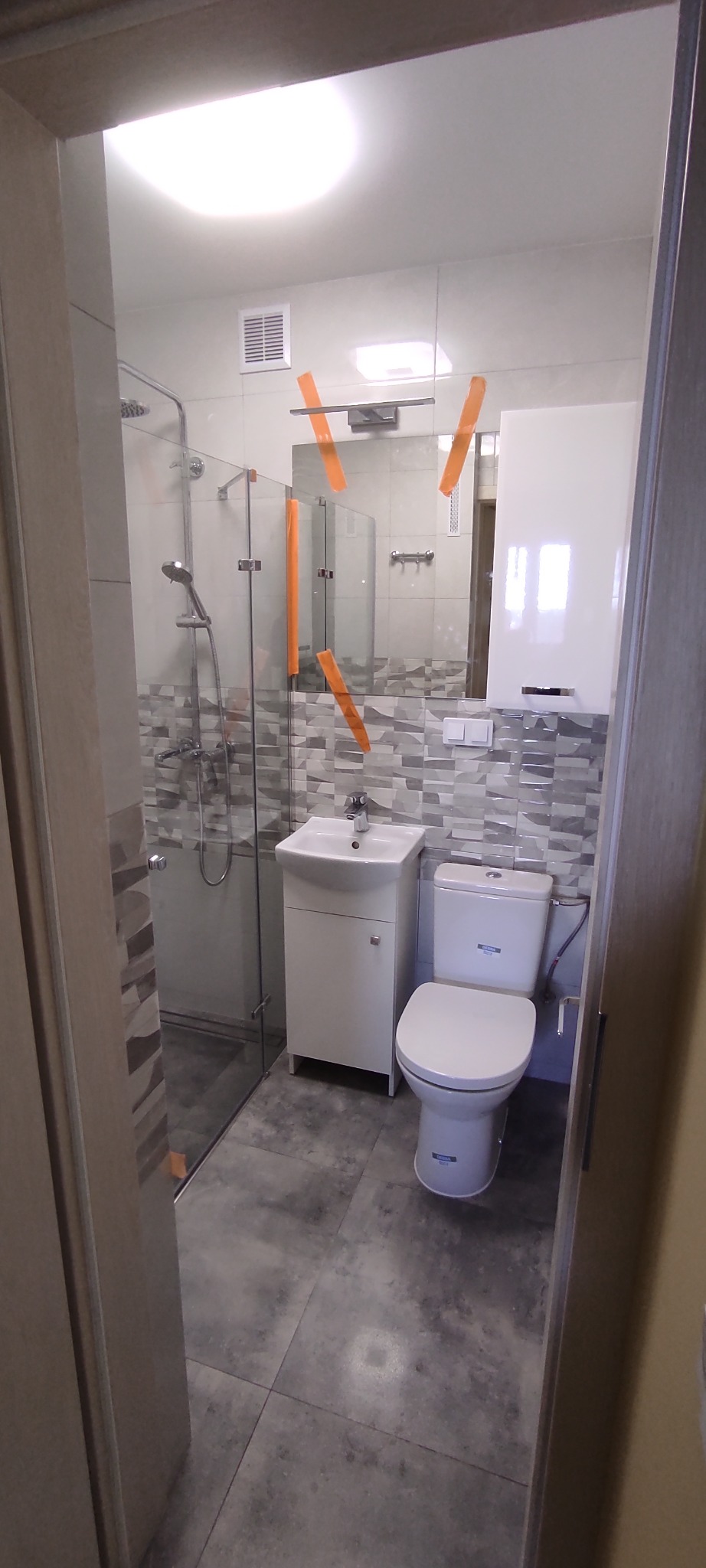 Nowoczesna łazienka z szarą mozaiką na ścianach, kabiną prysznicową ze szklanymi drzwiami, białą szafką pod umywalką i toaletą. Pomarańczowe paski zabezpieczające na lustrze i oświetleniu.
