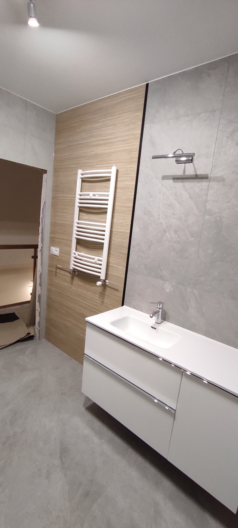 Wykończona łazienka z szarą marmurową płytką, drewnopodobną ścianą, białym grzejnikiem drabinkowym i szafką pod umywalkę z chromowanymi uchwytami, widok na niedokończone wejście do pomieszczenia obok.