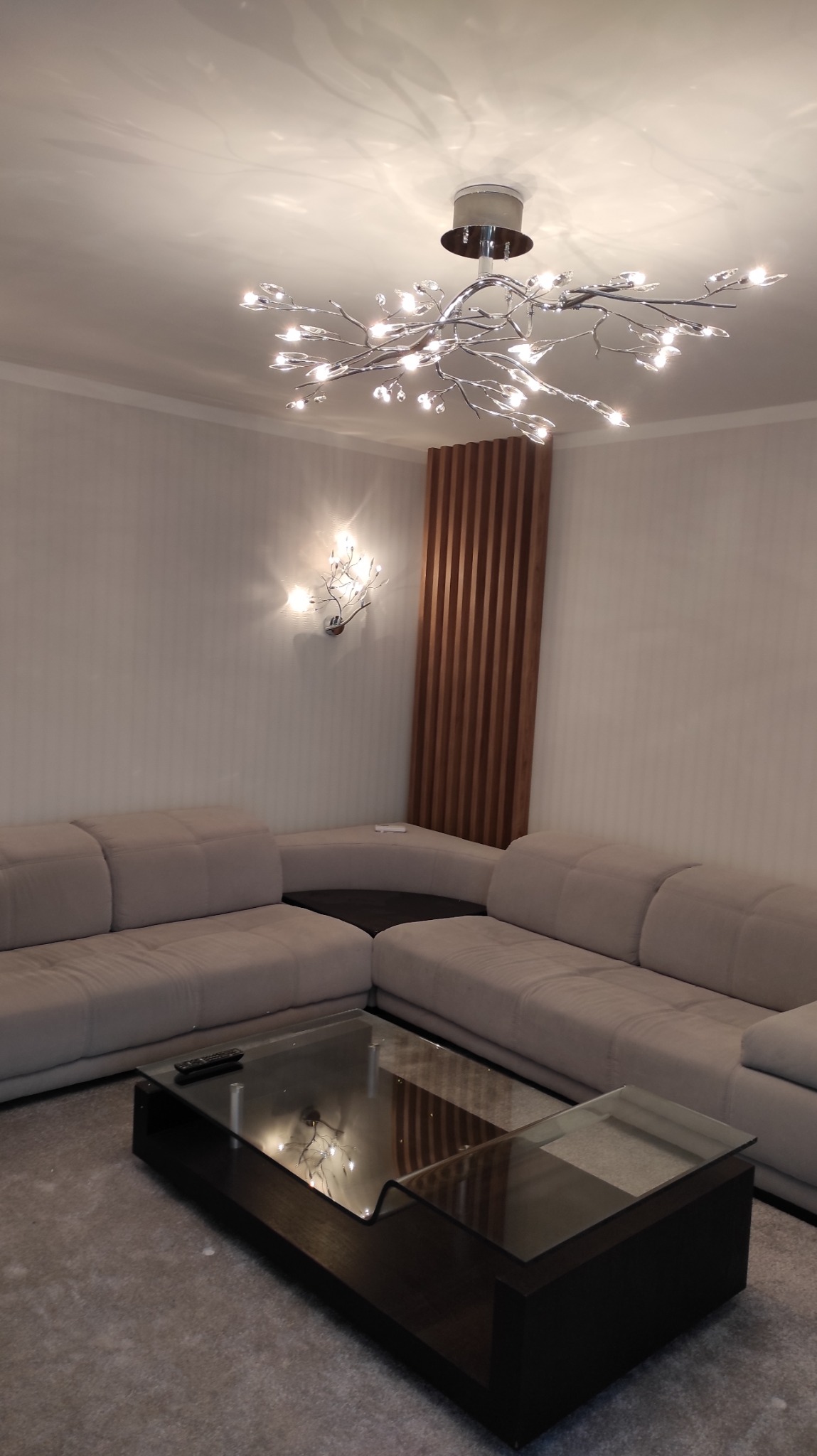 Jasny salon z narożną sofą, szklanym stolikiem kawowym i designerską lampą sufitową w kształcie gałęzi z kryształkami, oświetlona drewniana ścianka dekoracyjna w tle.