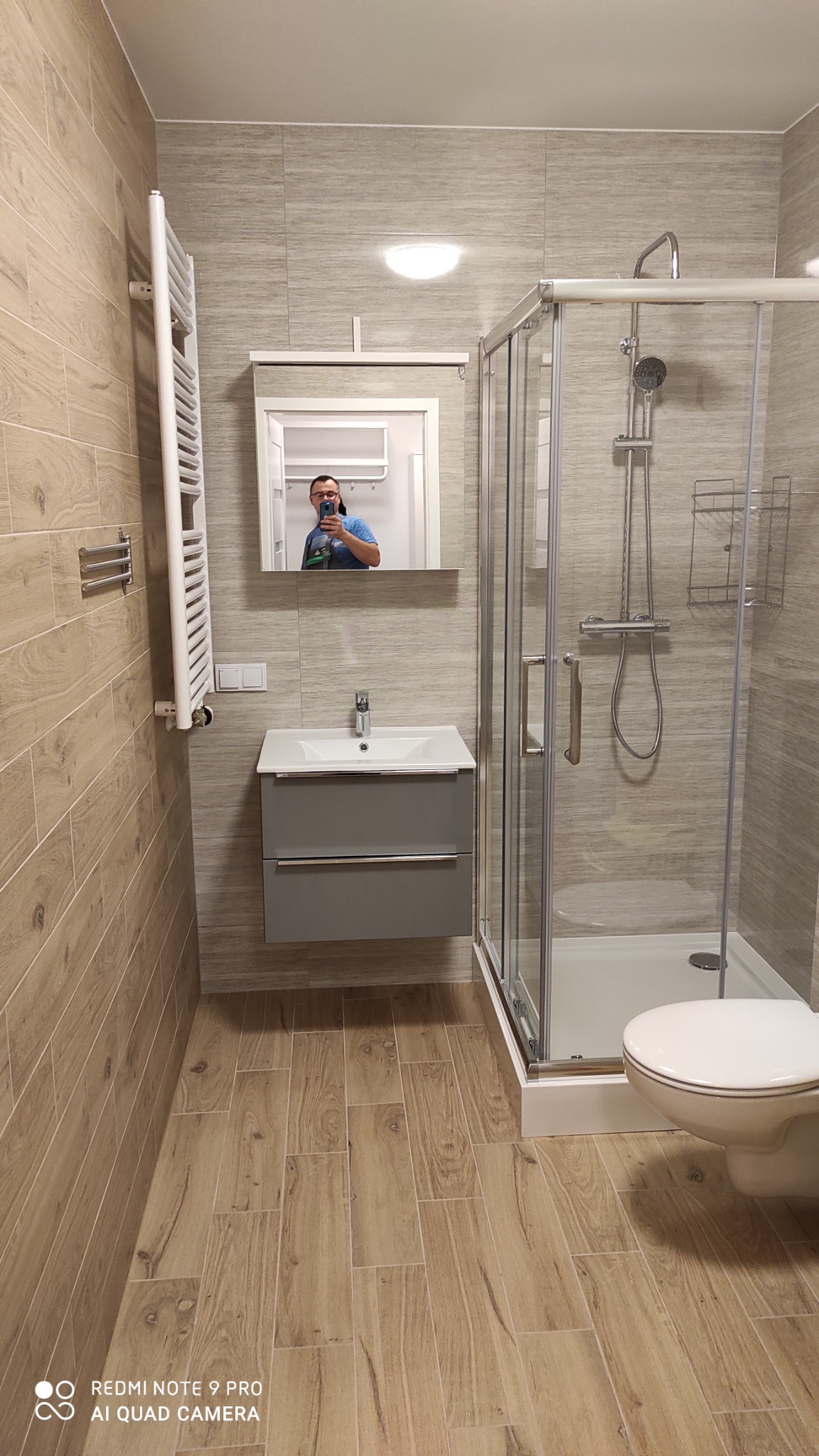 Nowoczesna łazienka z kabiną prysznicową, umywalką z szarą szafką i grzejnikiem drabinkowym, wykończona płytkami imitującymi drewno. W lustrze odbija się osoba robiąca zdjęcie.