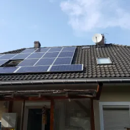 Instalacja fotowoltaiczna na dachu domu z brązową dachówką, widoczny otwarty lukarn, antena satelitarna i fragment zadaszonego tarasu.