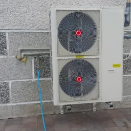 Zewnętrzna jednostka pompy ciepła zamontowana na szarej, teksturowanej ścianie budynku, widoczne rury i niebieski wąż doprowadzający wodę.