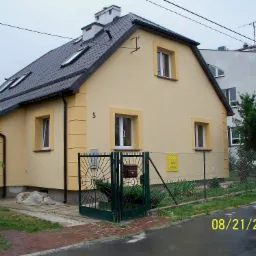 Dwukondygnacyjny dom z żółtą elewacją, ciemną dachówką i białymi ramami okiennymi, numer 5 na ścianie oraz zielona metalowa brama z dekoracyjnymi elementami.