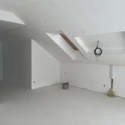 Poddasze po pracach wykończeniowych, ściany i sufit pomalowane na biało, widoczne okno dachowe, wystające przewody elektryczne, puszka z farbą i instalacja elektryczna w listwie.