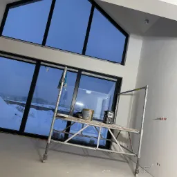 Wnętrze w trakcie szpachlowania ścian, widoczne rusztowanie z wiadrem farby i narzędziami, duże okna z widokiem na zimowy krajobraz.