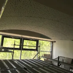 Montaż sufitu podwieszanego z perforowanych paneli na stelażu metalowym, widok z rusztowania na tle okna z zielenią na zewnątrz.