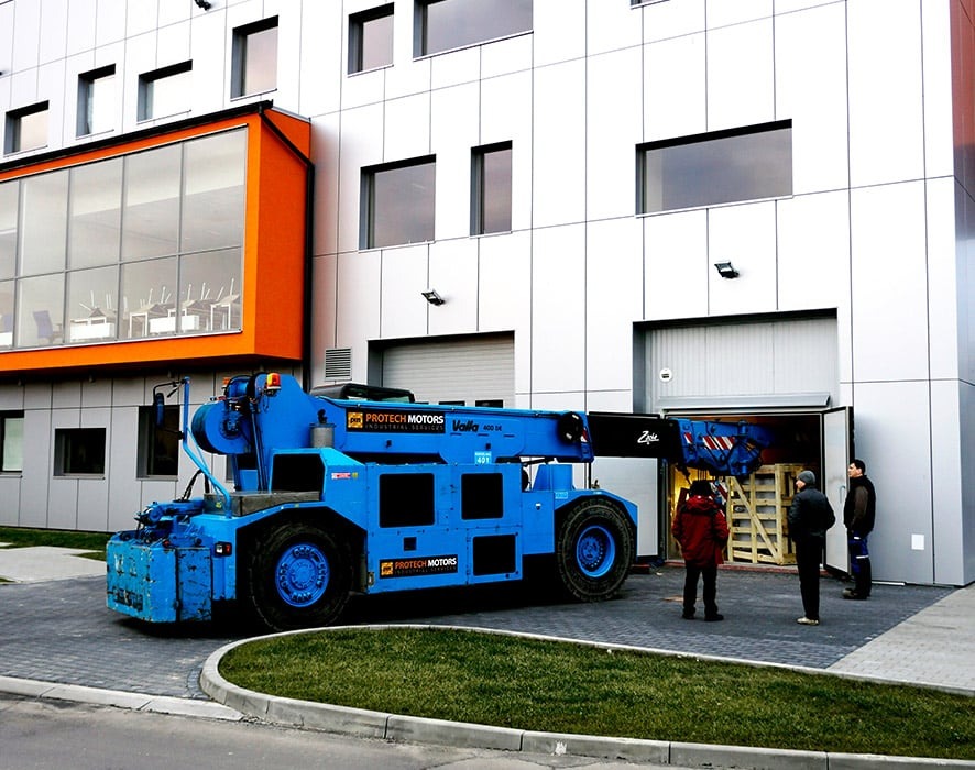 Niebieski samojezdny żuraw Valla 400 DC z wysuniętym ramieniem, ustawiony przed budynkiem z otwartą bramą garażową, gdzie widoczne są drewniane skrzynie, oraz stojący obok trzej mężczyźni.