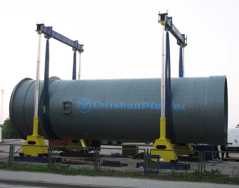 Ciężki, cylindryczny element konstrukcyjny z logo 'Christian Pfeifer' podnoszony za pomocą specjalistycznych dźwigów z żółtymi podstawami, widoczne otwory wentylacyjne i metalowe liny.