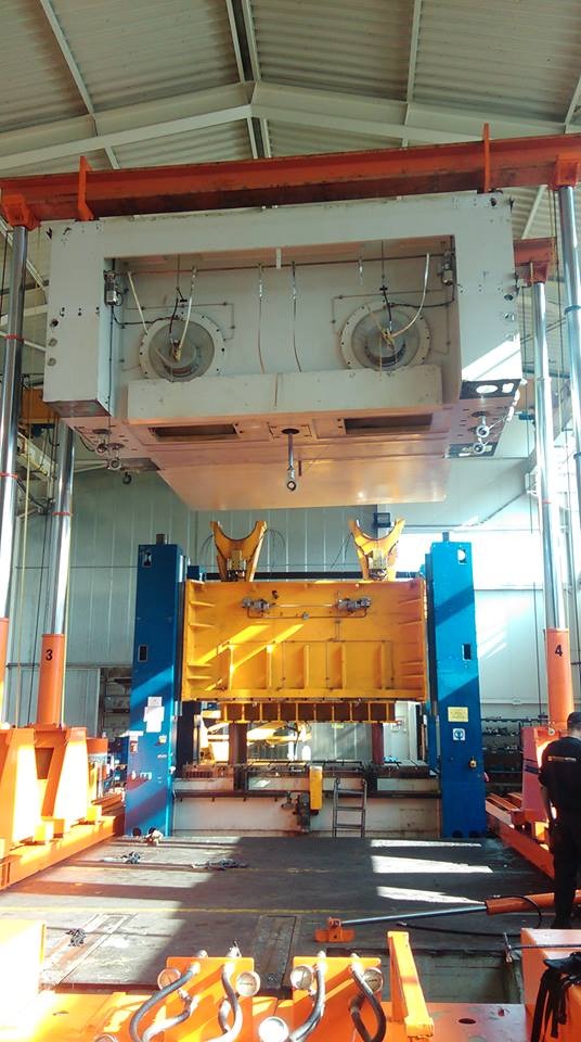 Wielkogabarytowa prasa przemysłowa w hali produkcyjnej, z widocznymi elementami konstrukcyjnymi w kolorach pomarańczowym, niebieskim i żółtym.