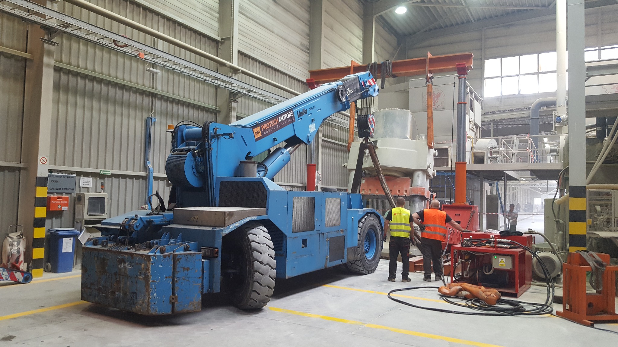 Niebieski dźwig samojezdny Protech Motors podnosi element maszyny w hali przemysłowej, pracownicy w kamizelkach odblaskowych nadzorują operację, widoczne okablowanie i urządzenia pomocnicze.
