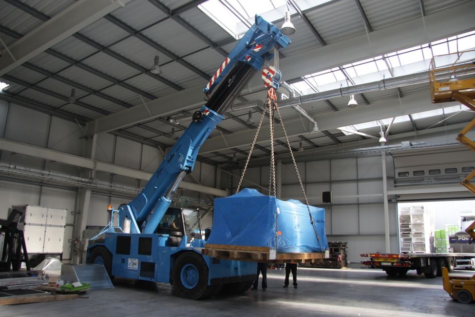 Niebieski dźwig samojezdny podnosi zabezpieczoną folią paletę w hali magazynowej z oknami dachowymi.