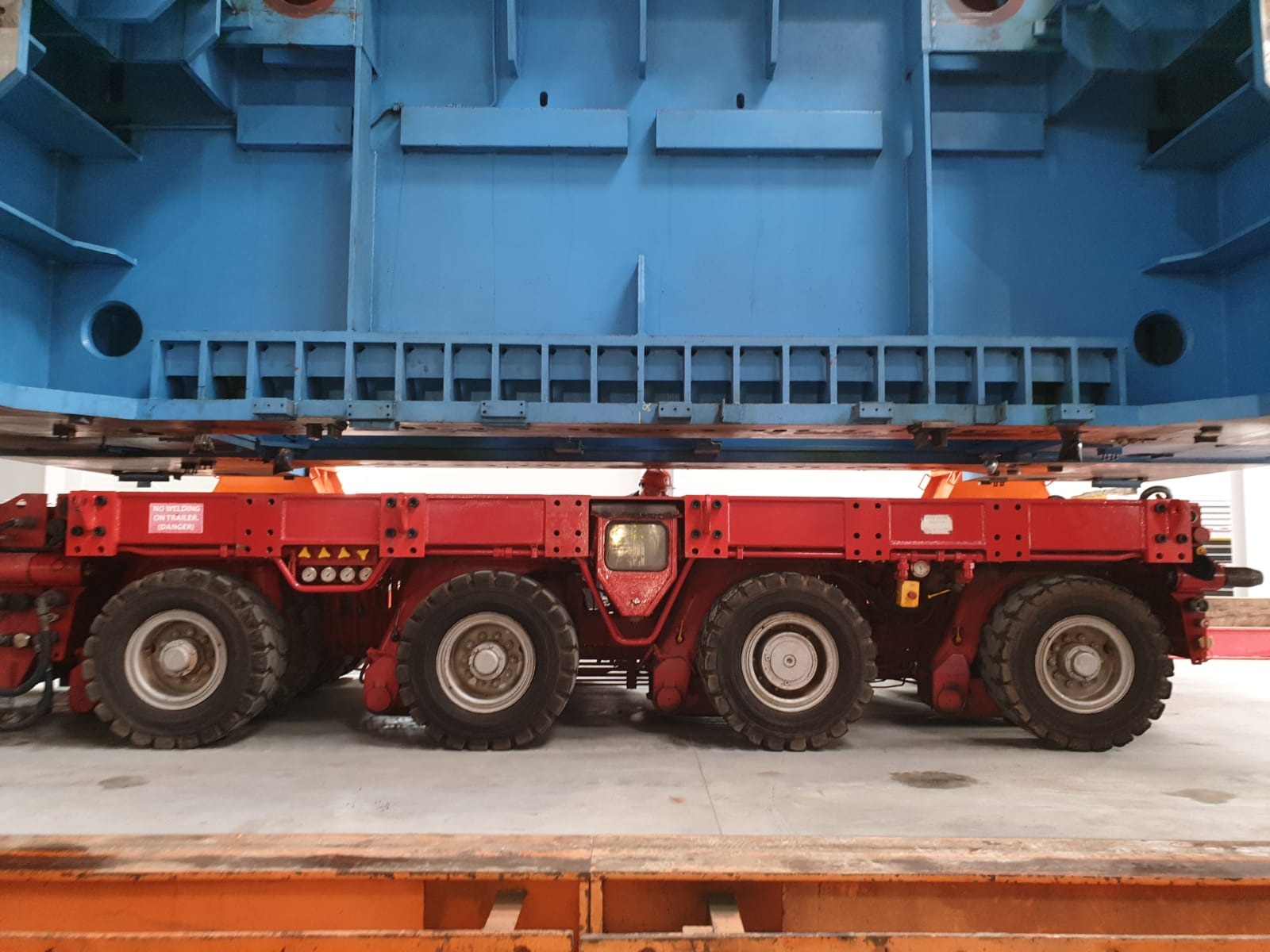 Czerwony, wielokołowy transporter modułowy (SPMT) przewożący niebieski, wielkogabarytowy element konstrukcyjny w hali.