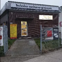 Fasada budynku firmy oferującej technikę sanitarno-grzewczą, widoczny szyld z nazwą i numerem telefonu, tablica informacyjna oraz reklamy produktów na zewnątrz.