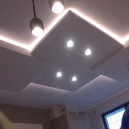 Arek Bancer - Wielopoziomowy sufit podwieszany z oświetleniem LED i wpuszczanymi reflektorami, widoczne wiszące lampy i fragment ściany z dekoracyjną cegłą.