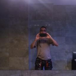 Arek Bancer - Mężczyzna w okularach robi telefonem zdjęcie w lustrze łazienki z szarymi płytkami i oświetleniem LED nad wanną.