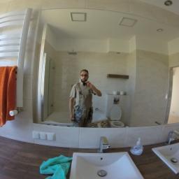 Arek Bancer - Odbicie w lustrze wyremontowanej łazienki z beżowymi płytkami, białą ceramiką i brązowym blatem. W lustrze widoczny mężczyzna robiący zdjęcie telefonem.