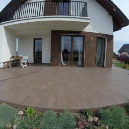 Arek Bancer - Szeroki kadr na duży, półokrągły taras wyłożony brązowymi płytkami imitującymi drewno, z widokiem na dwupiętrowy dom z drewnianą elewacją i balkonem, otoczony zielonym ogrodem z rabatami kwiatowymi.