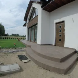 Nowoczesny dom jednorodzinny z tarasem wykończonym brązowymi płytkami imitującymi drewno, widoczne schody wejściowe i fragment ogrodu w trakcie aranżacji.