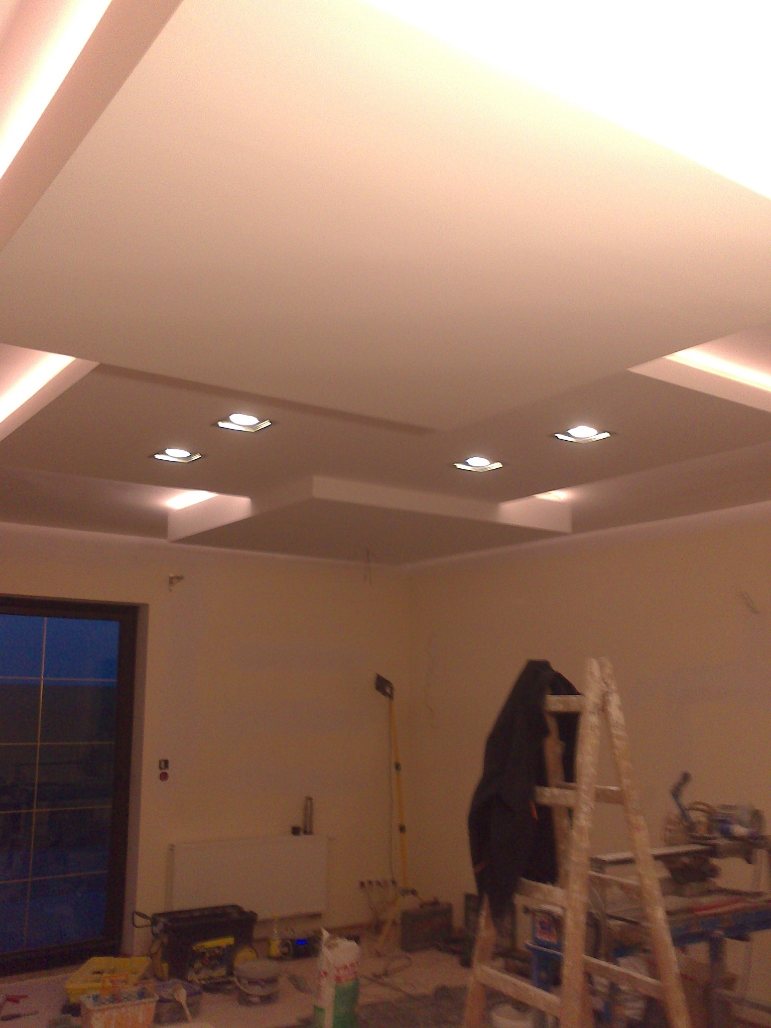 Wykończony sufit podwieszany z oświetleniem punktowym, widoczna drabina i narzędzia malarskie w tle, sugerujące trwające prace wykończeniowe.