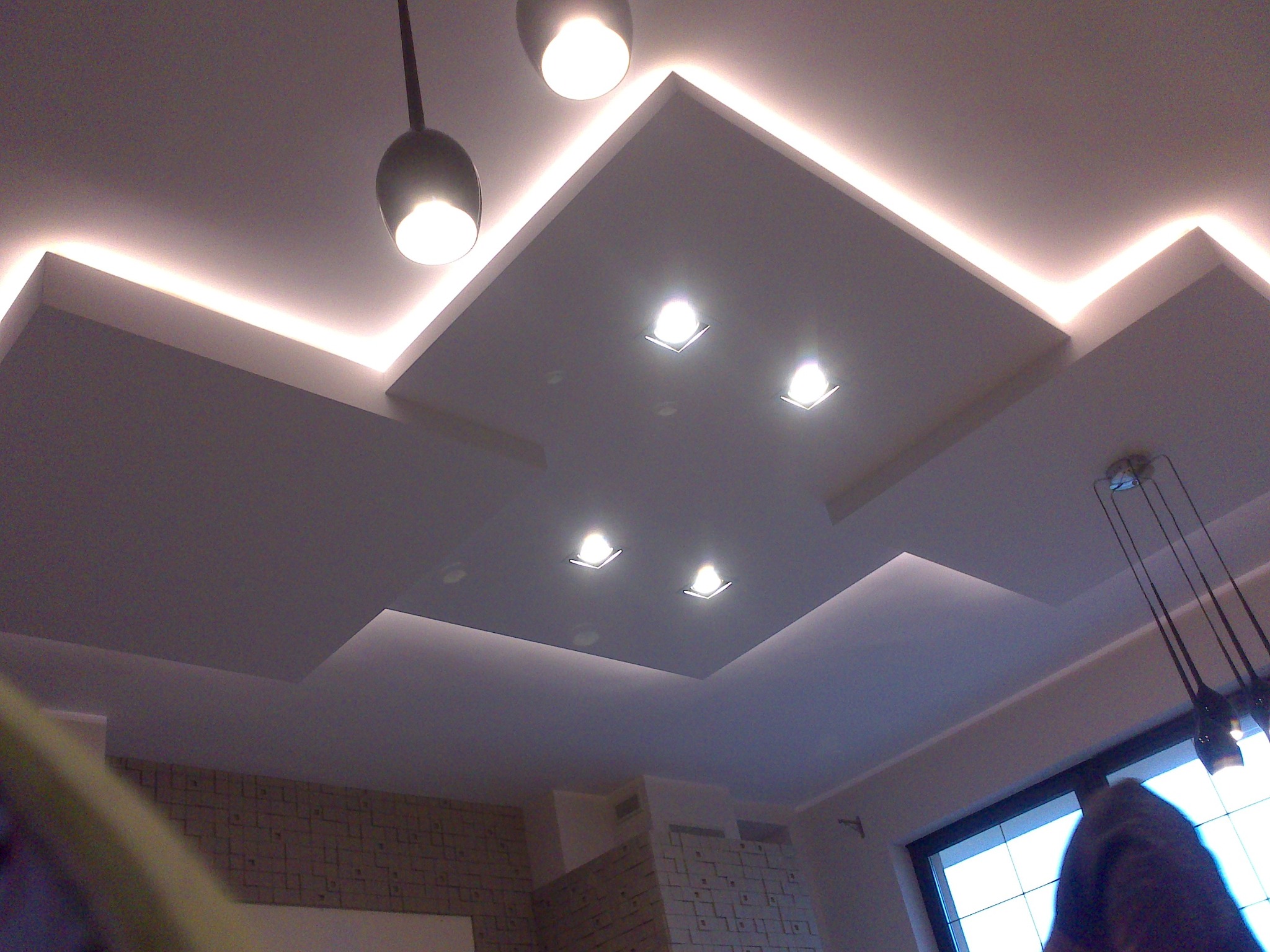 Wielopoziomowy sufit podwieszany z oświetleniem LED i wpuszczanymi reflektorami, widoczne wiszące lampy i fragment ściany z dekoracyjną cegłą.