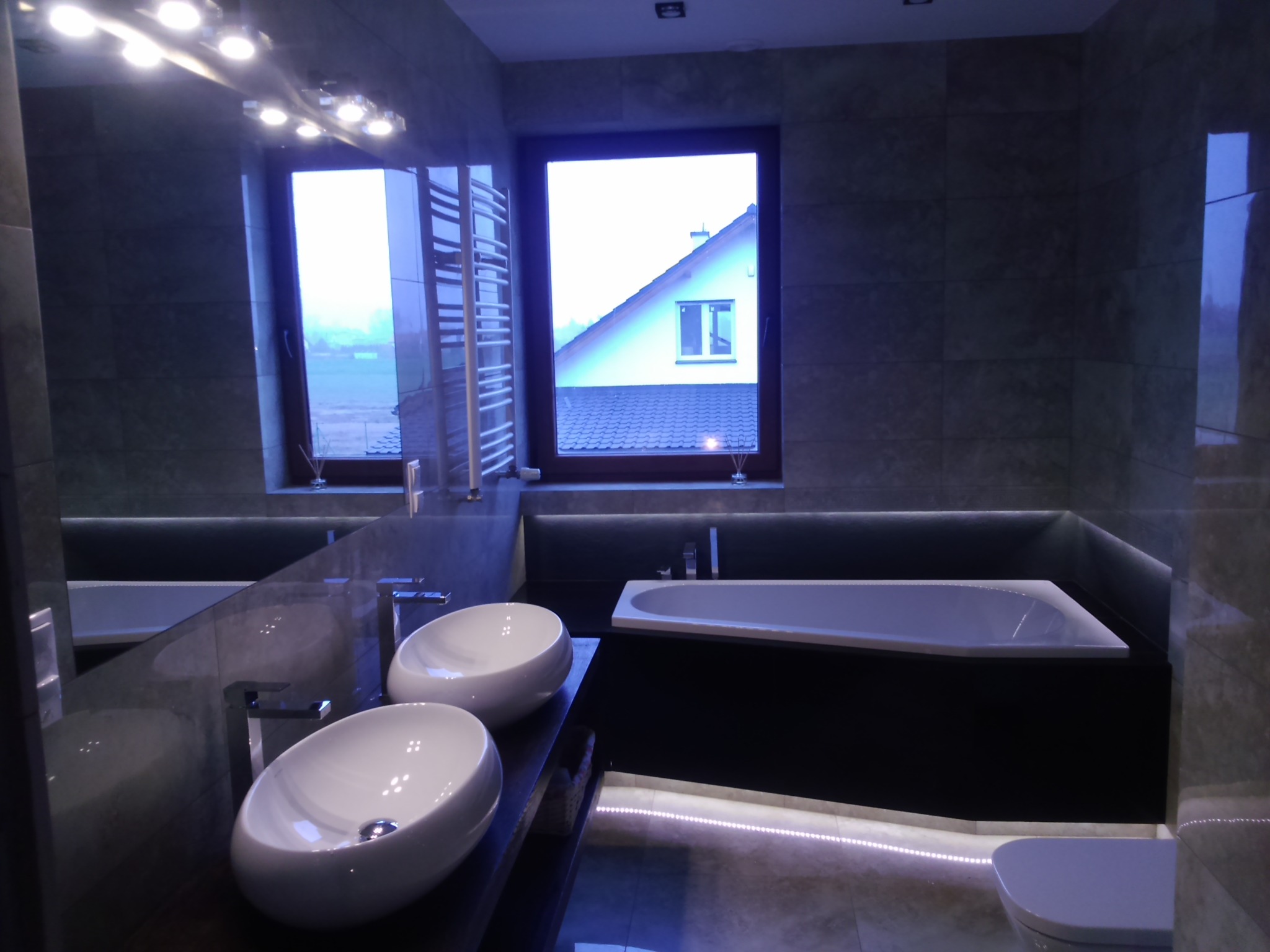 Nowoczesna łazienka w szarościach z podświetlaną wanną, dwiema owalnymi umywalkami nablatowymi i oknem z widokiem na dom.