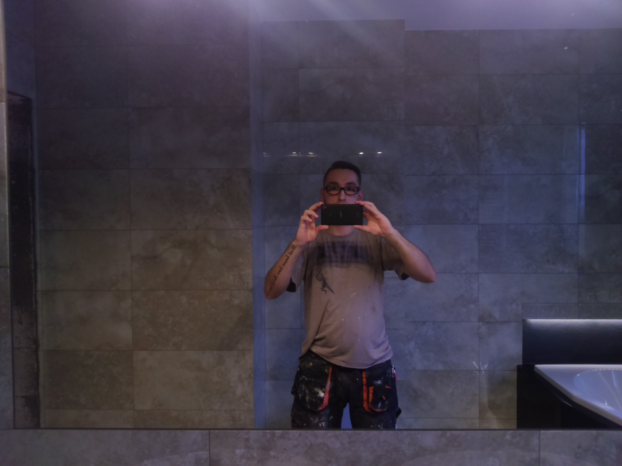 Mężczyzna w okularach robi telefonem zdjęcie w lustrze łazienki z szarymi płytkami i oświetleniem LED nad wanną.