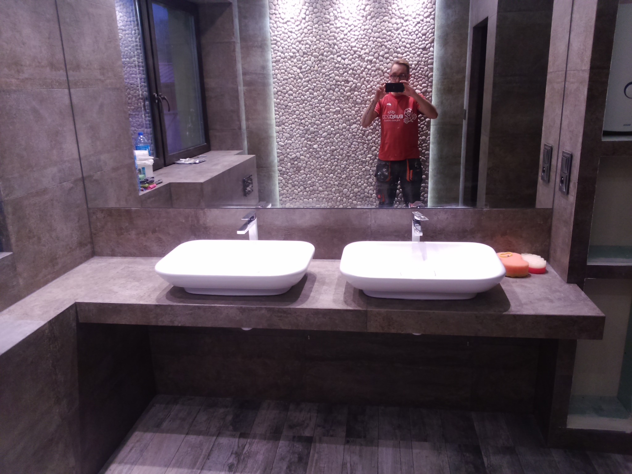 Nowoczesna łazienka z dwoma białymi umywalkami nablatowymi na blacie z ciemnego kamienia, ściana wyłożona dekoracyjnymi kamieniami, podłoga z paneli imitujących drewno. W lustrze odbija się osoba...