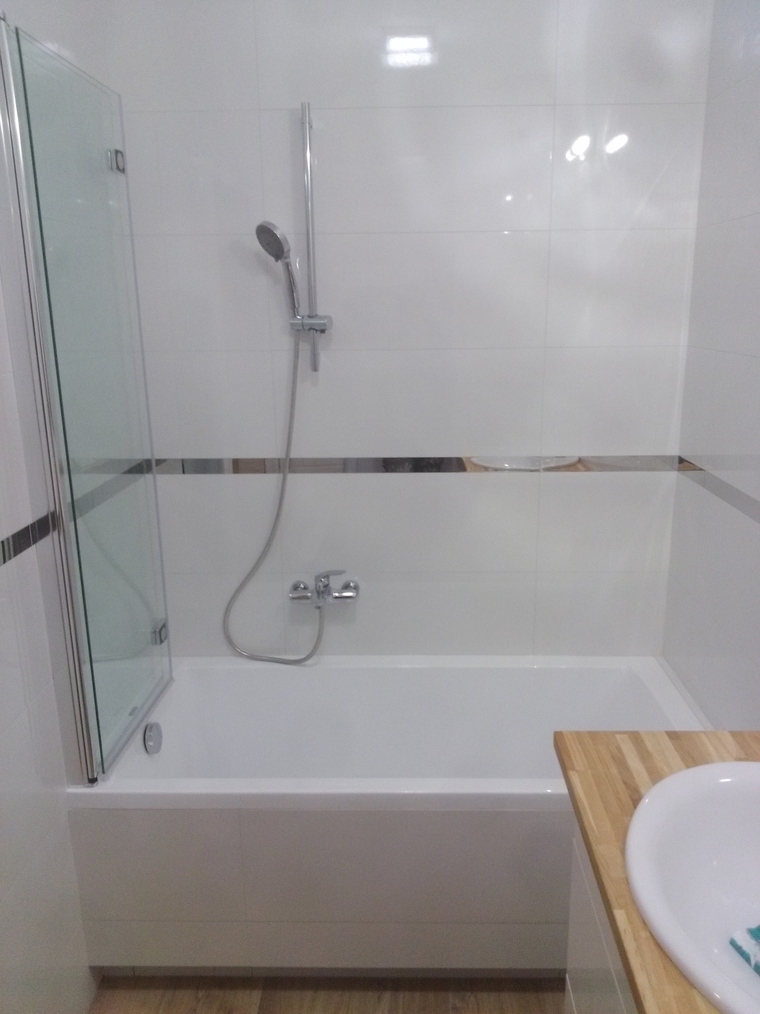 Biała łazienka w Toruniu po remoncie, z wanną, szklanym parawanem nawannowym i chromowaną armaturą, wykończona płytkami z dekoracyjnym pasem.