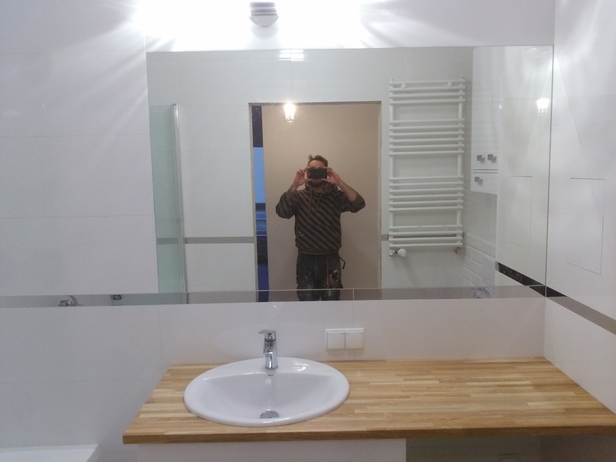 Nowoczesna łazienka z białą umywalką na drewnianym blacie, widoczny grzejnik drabinkowy i odbicie fotografa w lustrze.