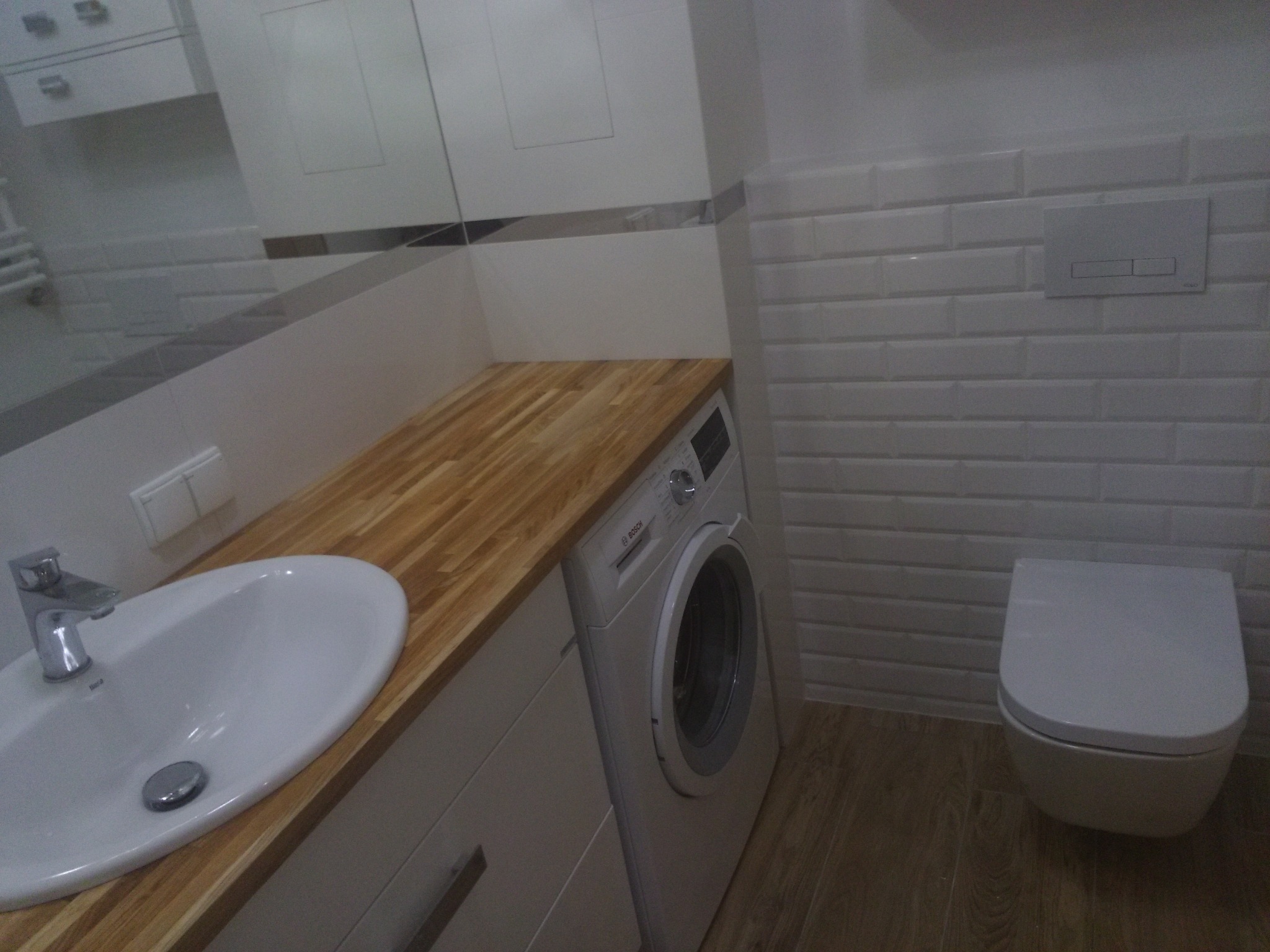 Nowoczesna łazienka z drewnianym blatem pod umywalką, białą zabudową meblową, pralką Bosch i podwieszaną toaletą na tle białych płytek imitujących cegłę.
