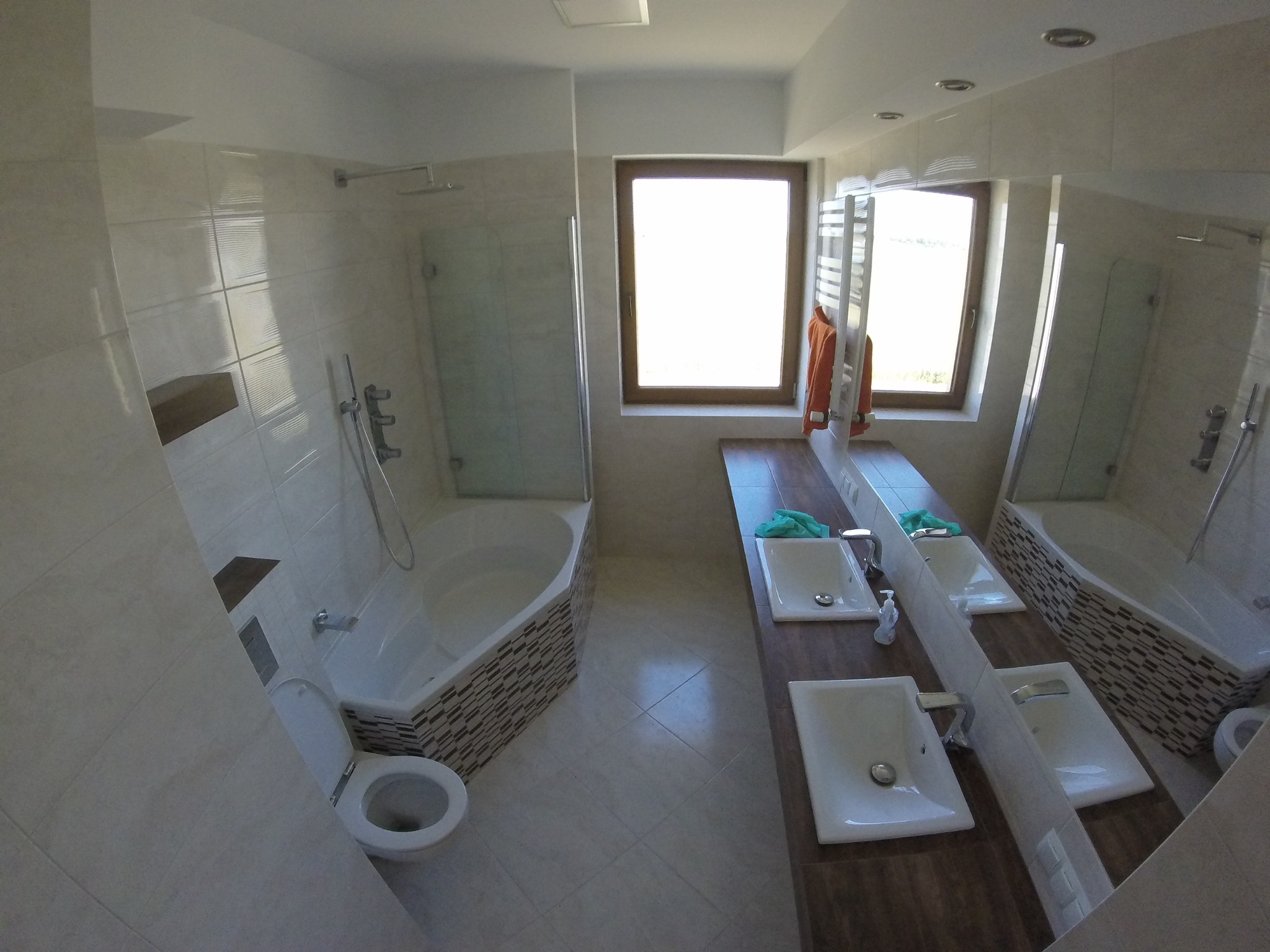 Jasna łazienka po remoncie z wanną obudowaną mozaiką, dwoma umywalkami na drewnianym blacie i oknem w tle.