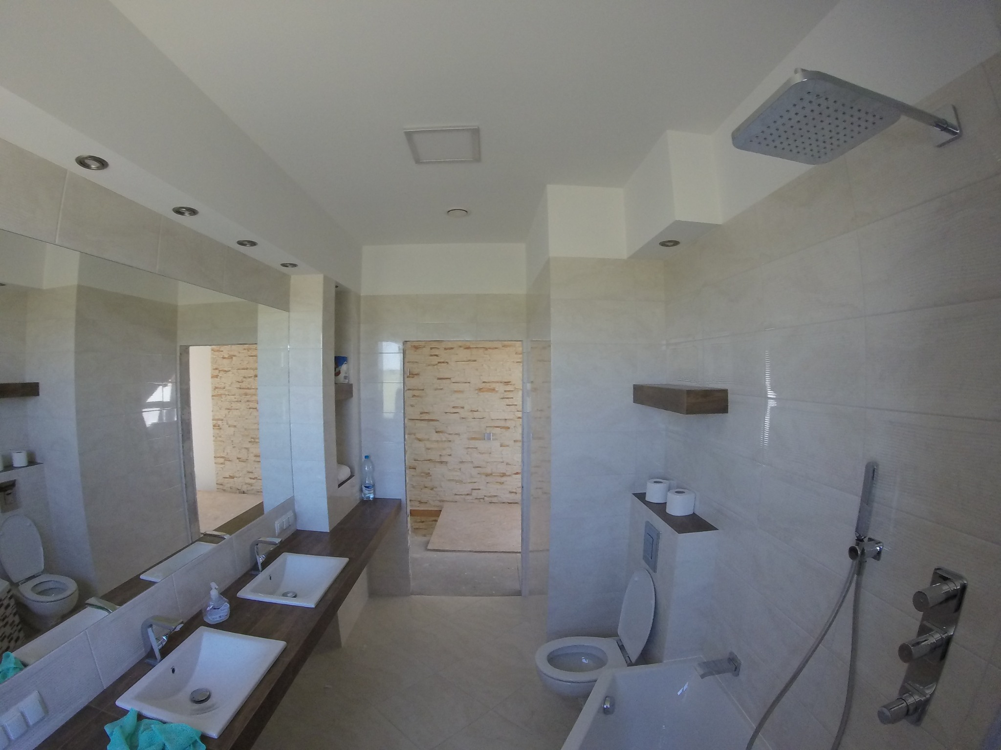 Jasna łazienka z dwoma umywalkami na drewnianym blacie, dużym lustrem, kabiną prysznicową typu walk-in i podwieszaną toaletą z półką na papier toaletowy. Widoczne oświetlenie punktowe w suficie.