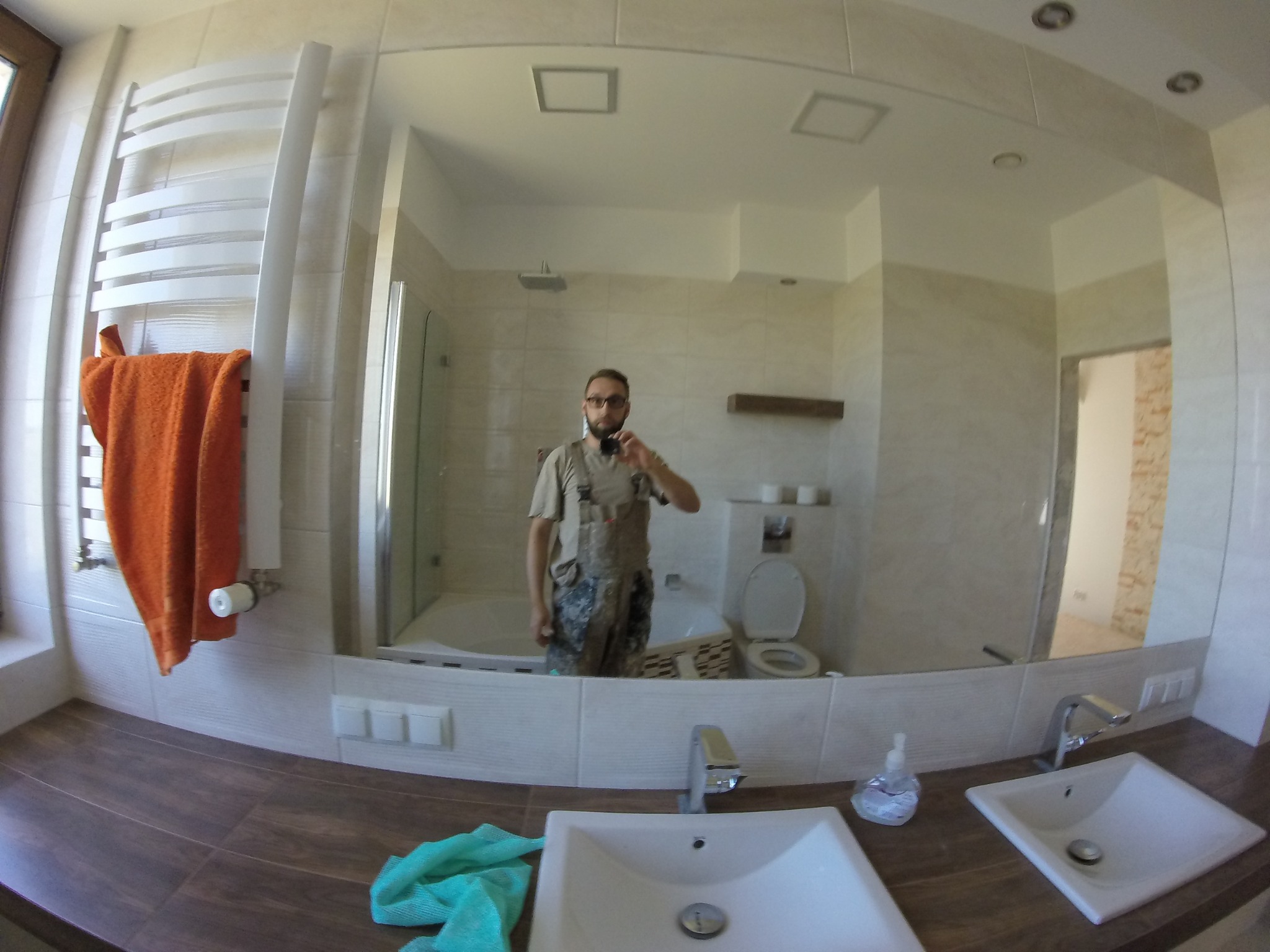 Odbicie w lustrze wyremontowanej łazienki z beżowymi płytkami, białą ceramiką i brązowym blatem. W lustrze widoczny mężczyzna robiący zdjęcie telefonem.