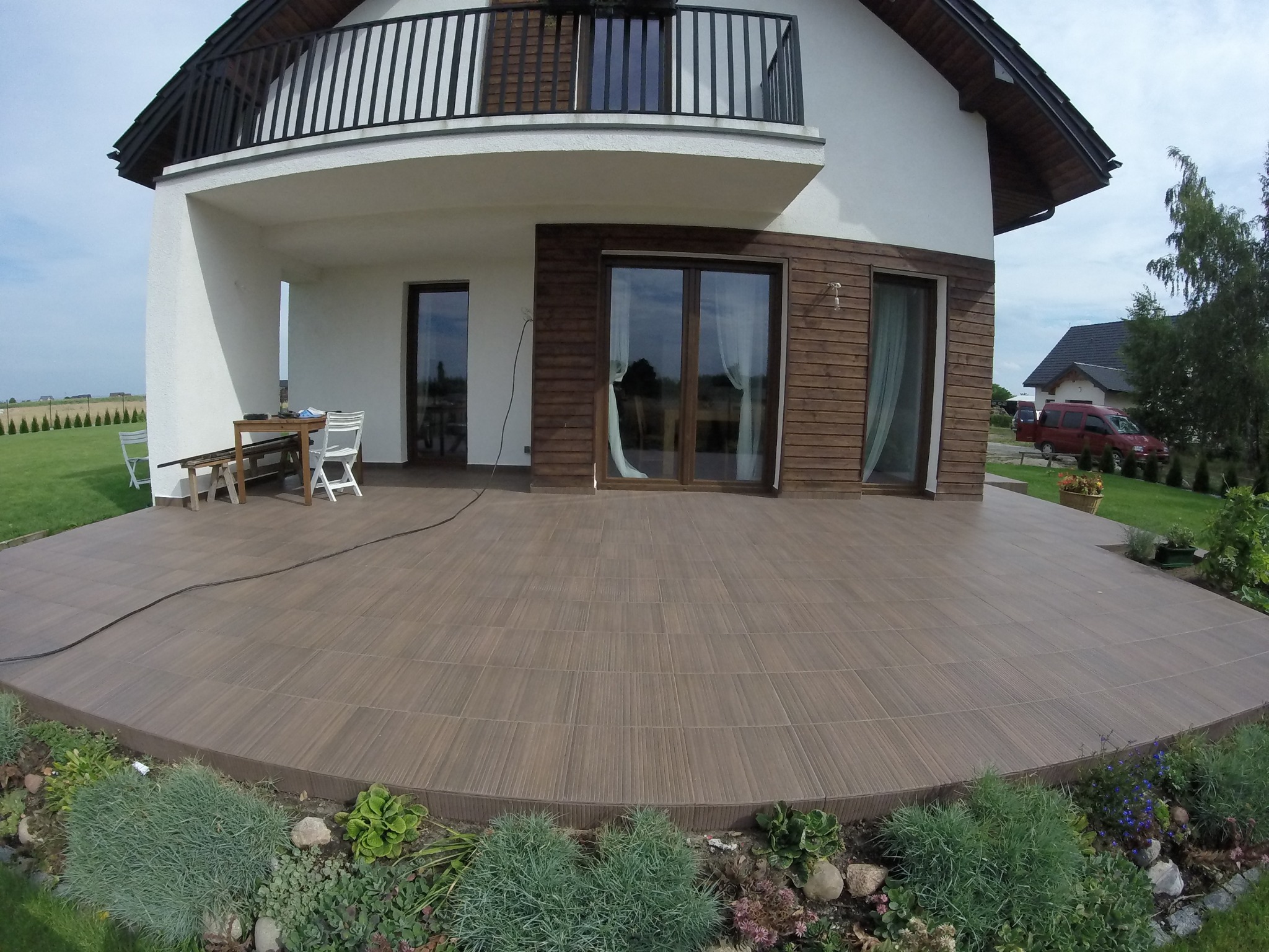 Szeroki kadr na duży, półokrągły taras wyłożony brązowymi płytkami imitującymi drewno, z widokiem na dwupiętrowy dom z drewnianą elewacją i balkonem, otoczony zielonym ogrodem z rabatami kwiatowymi.