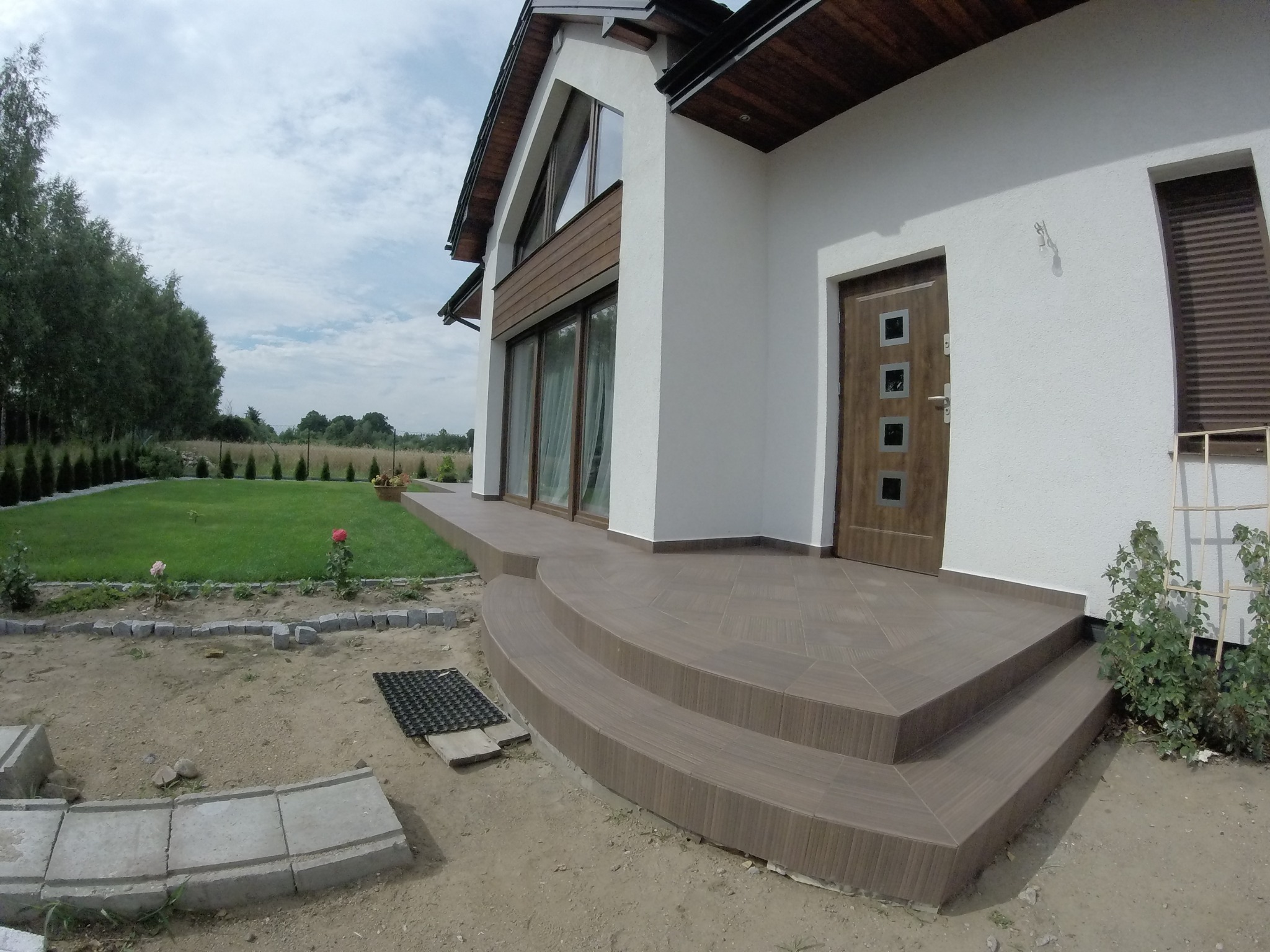 Nowoczesny dom jednorodzinny z tarasem wykończonym brązowymi płytkami imitującymi drewno, widoczne schody wejściowe i fragment ogrodu w trakcie aranżacji.