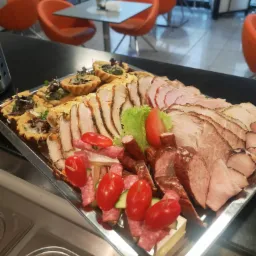 Srebrna taca z tartaletkami z ziołami, plastrami pieczonego mięsa, krojonej wędliny i koreczkami z pomidorków koktajlowych, salami i sera, na tle nowoczesnej sali z pomarańczowymi krzesłami.