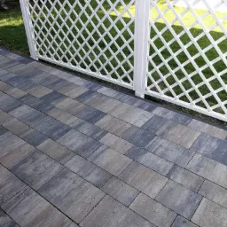 Ułożona nawierzchnia z kostki brukowej w odcieniach szarości i grafitu, przylegająca do białej, ażurowej ścianki działowej, z widocznym trawnikiem w tle.