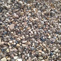 Nierównomierny stos różnokolorowych kamieni rzecznych, głównie w odcieniach beżu, brązu i szarości, o zróżnicowanych kształtach i wielkościach, oświetlonych naturalnym światłem słonecznym.