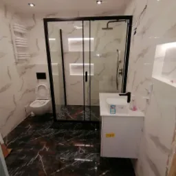 Nowoczesna łazienka z czarną kabiną prysznicową, marmurowymi płytkami na ścianach i podłodze w odcieniach brązu i czerni, białą szafką pod umywalką i podświetlanymi wnękami.