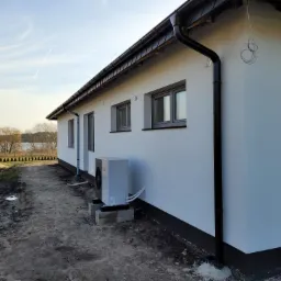 Biały, parterowy dom z szarą stolarką okienną i zamontowaną pompą ciepła na zewnątrz, widok z boku na tle jeziora i błękitnego nieba.