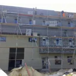 Wielopiętrowy budynek w trakcie ocieplania, widoczne rusztowania, częściowo ocieplona elewacja oraz pracownik w pomarańczowym kombinezonie na górnym piętrze.