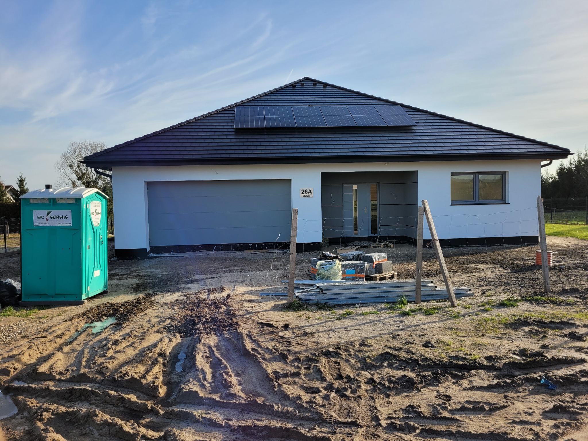 Nowoczesny parterowy dom z panelami słonecznymi na dachu, widoczna toaleta przenośna i materiały budowlane na błotnistym terenie.