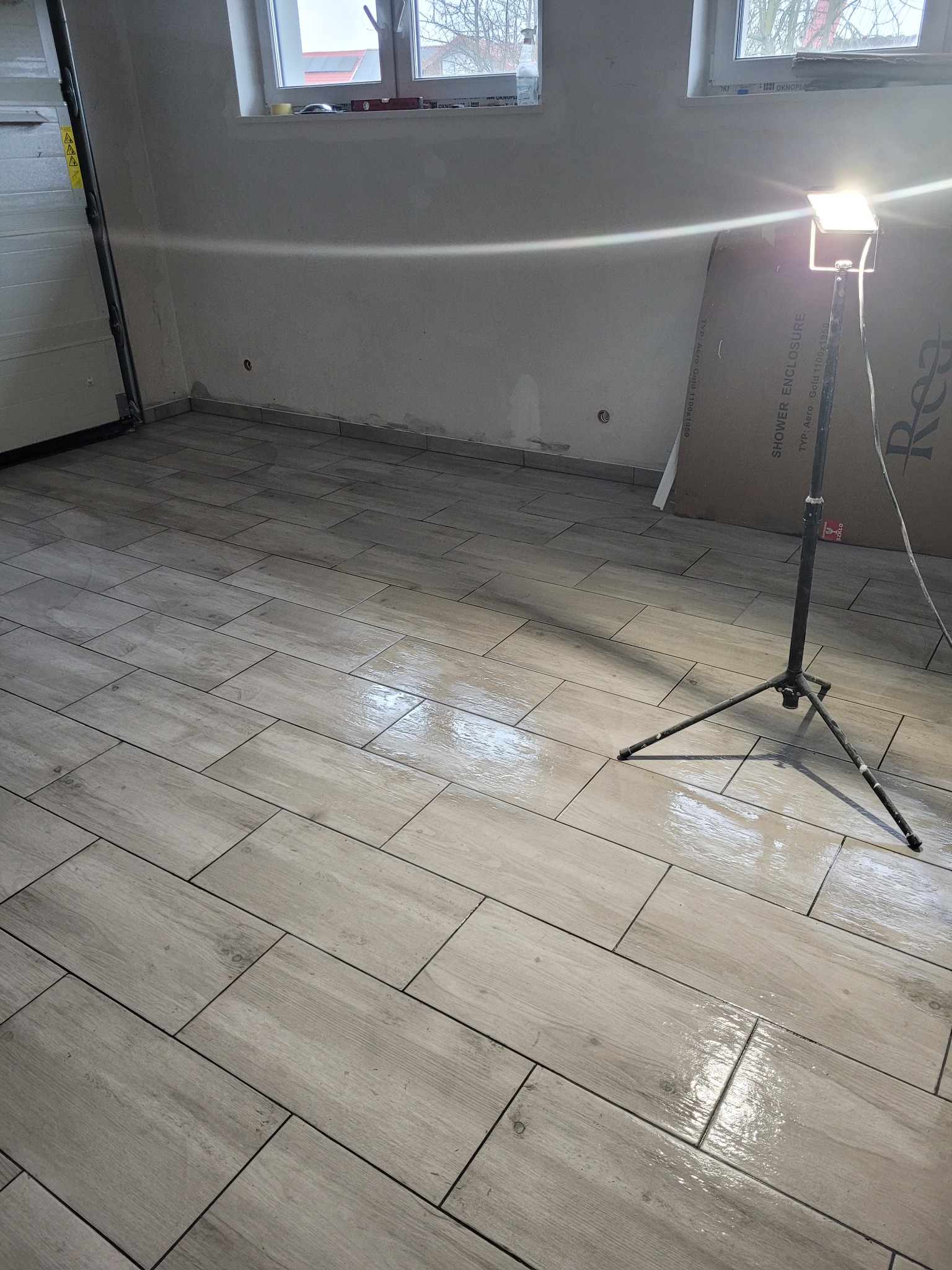 Ułożone płytki podłogowe imitujące drewno w jasnym odcieniu, oświetlone lampą budowlaną na trójnogu, widoczne okna i fragment bramy garażowej w tle.
